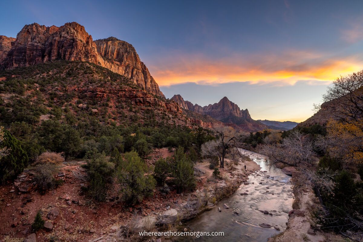 Photo of a curving river cutting through a red rock landscape with a tall formation to one side and colors in the sky at Canyon Junction Bridge in Zion during the winter