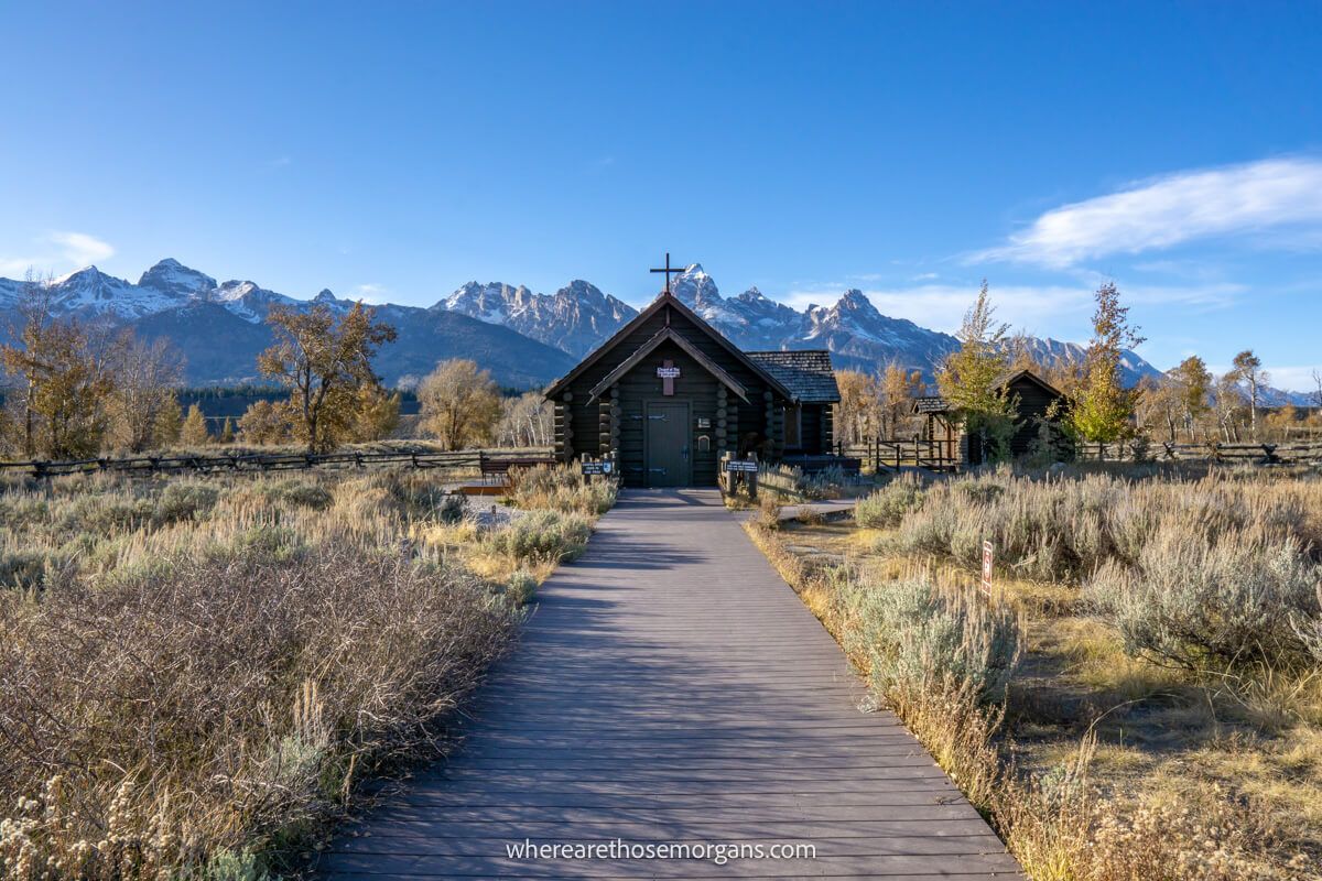 Wooden path leading through long golden grass to a small wooden church backed by snow capped mountains in Grand Teton National Park with a blue sky above