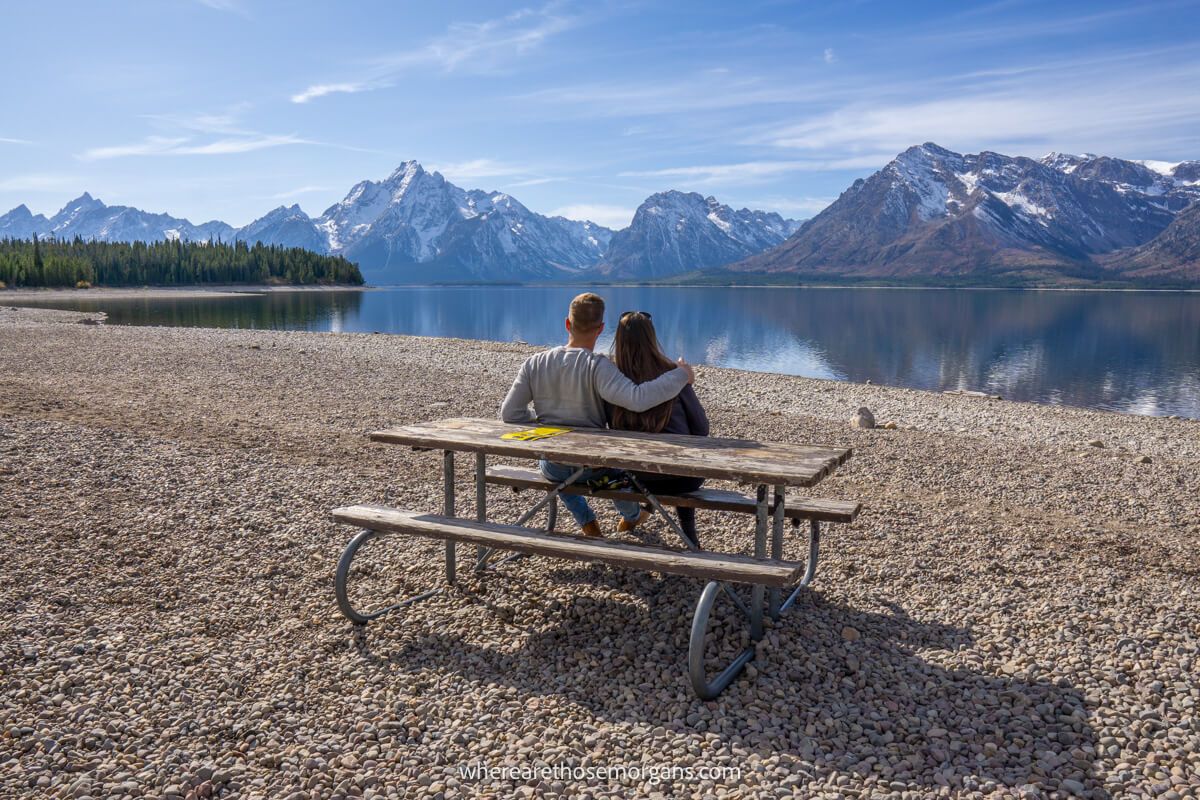 Couple sat at a bench on a rocky pebble beach looking at a view of a lake and mountains in Grand Teton