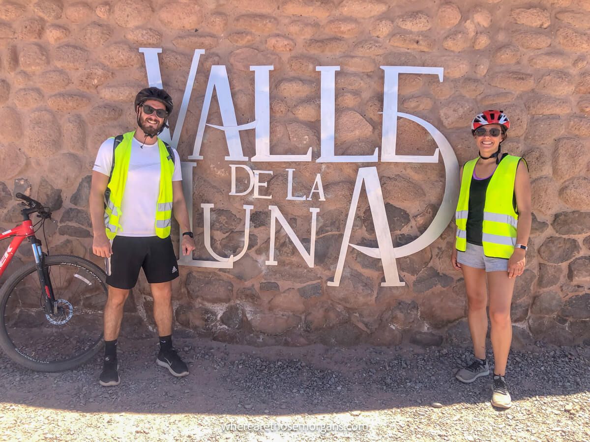 Photo of a couple in cycling gear standing on either side of a Valle de la Luna sign stuck to a brick wall with bikes to the side