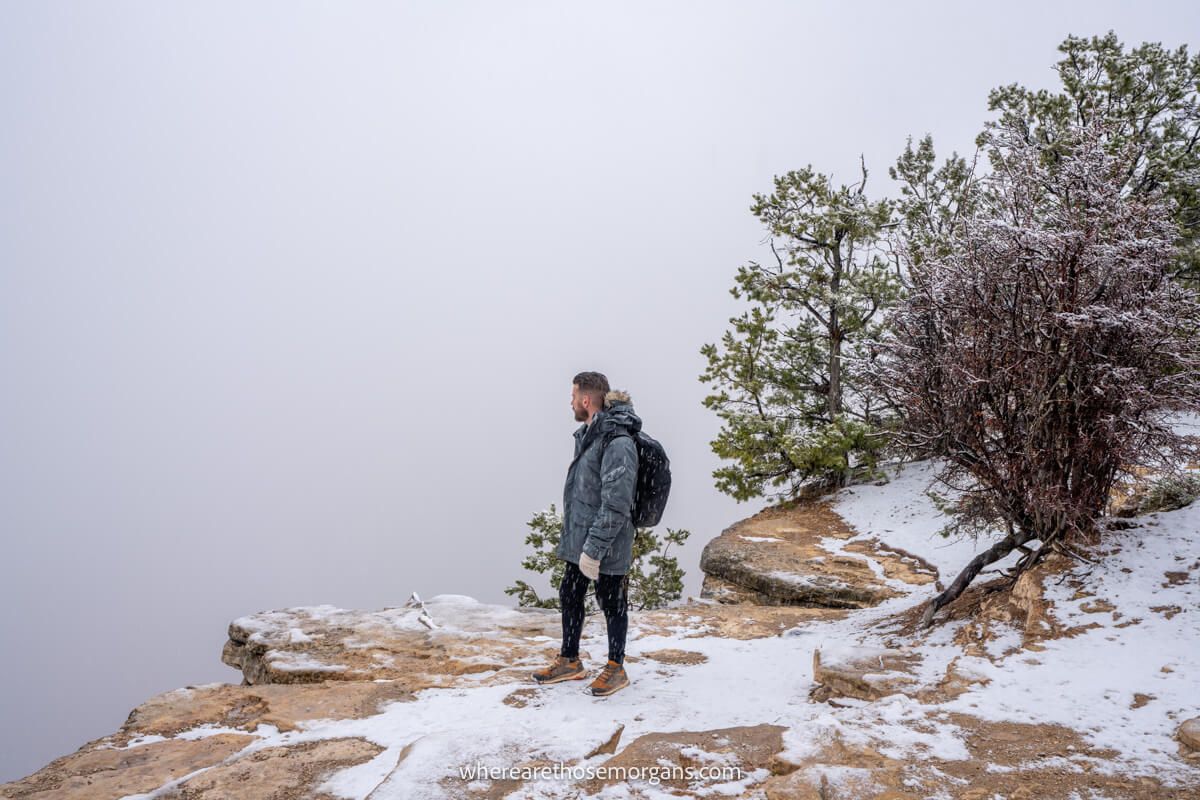 Tourist in heavy winter coat standing on snowy rocks looking out at a vast landscape filled with thick dense fog and snow clouds