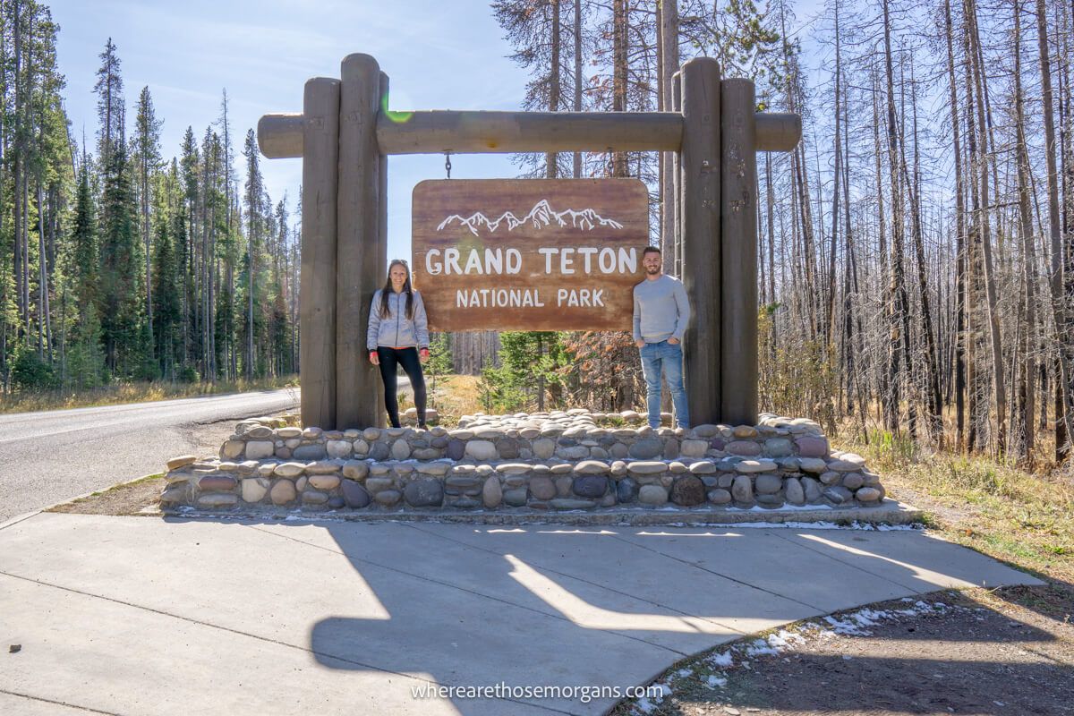 Couple standing on either side of the Grand Teton National Park entrance sign on a sunny day