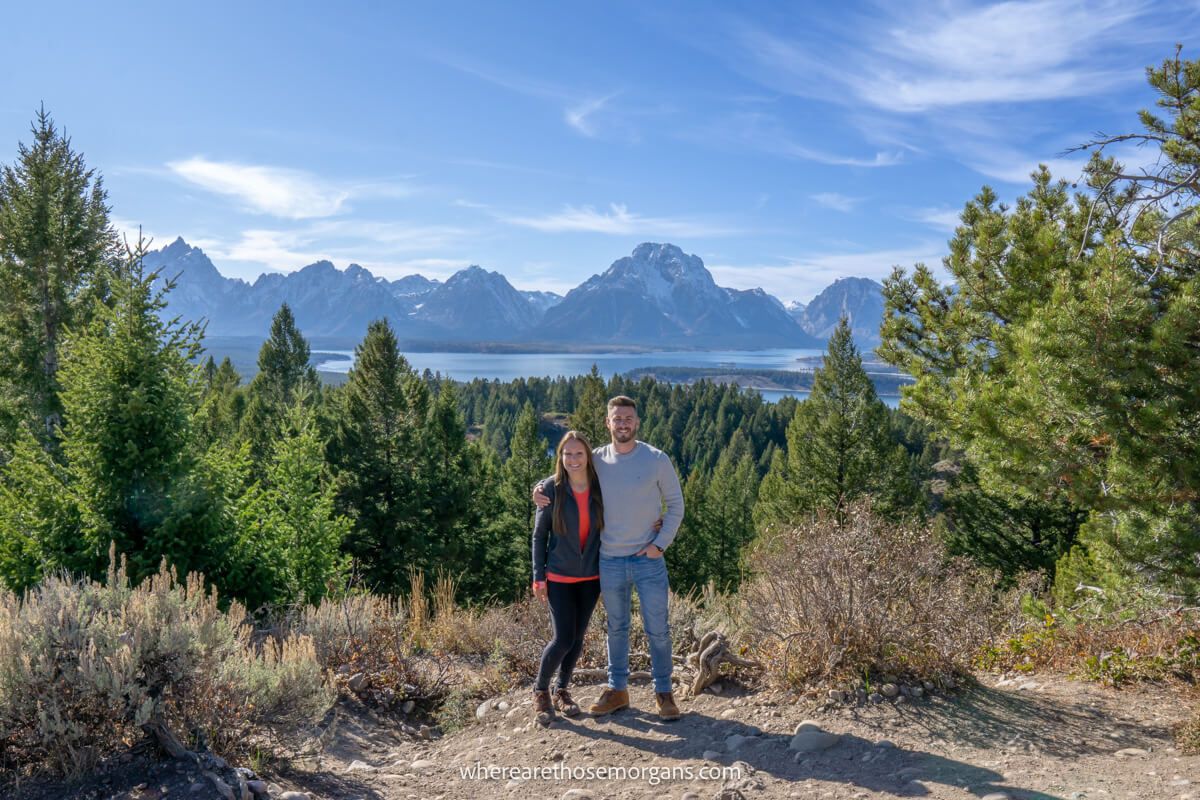 Couple standing together for a photo at an elevated vantage point overlooking evergreen trees, a lake and distant mountains on a clear sunny day in Grand Teton National Park