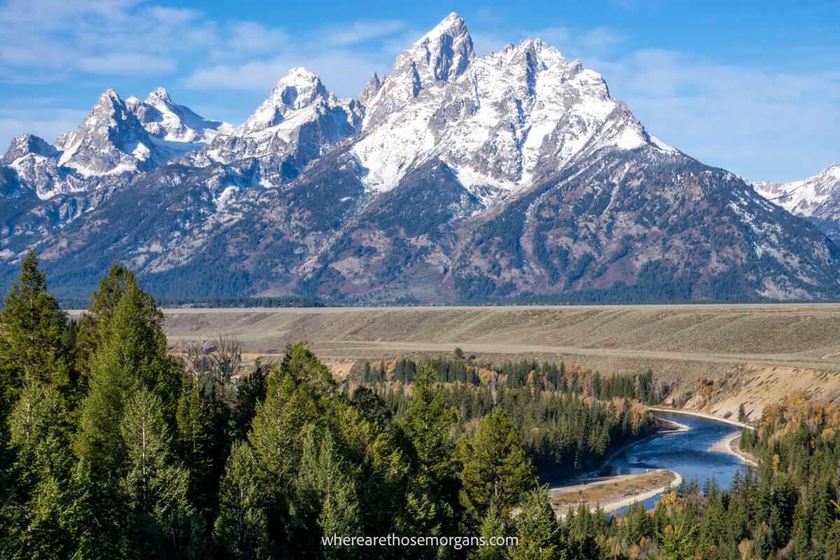 Tall snow capped mountains bursting into the sky from a flat meadow with a river curving in the foreground