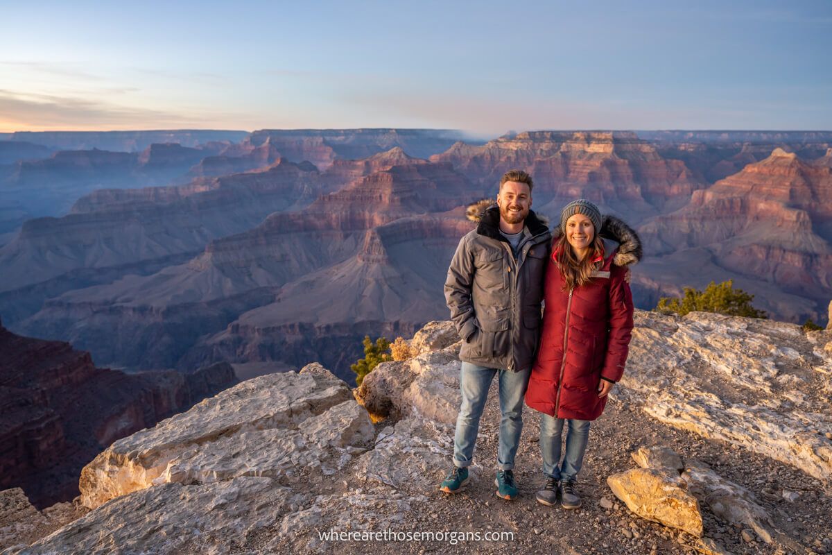 Photo of a couple standing together for a photo in winter coats on a rocky surface with views over Grand Canyon South Rim behind at sunset