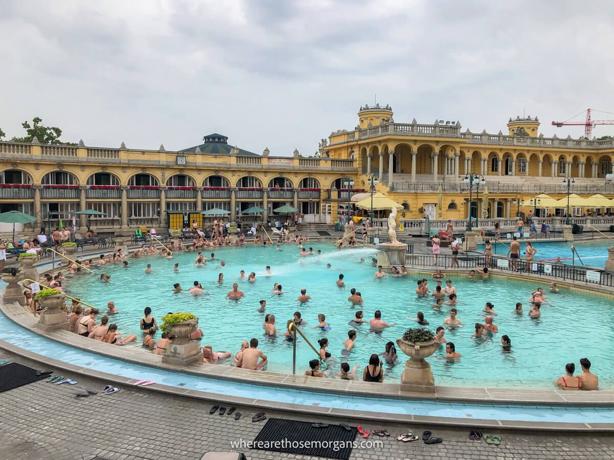 Photo of a large outdoor thermal pool packed full of tourists backed by a long yellow building with old world architecture under a cloudy sky at Szechenyi Baths in Budapest, Hungary