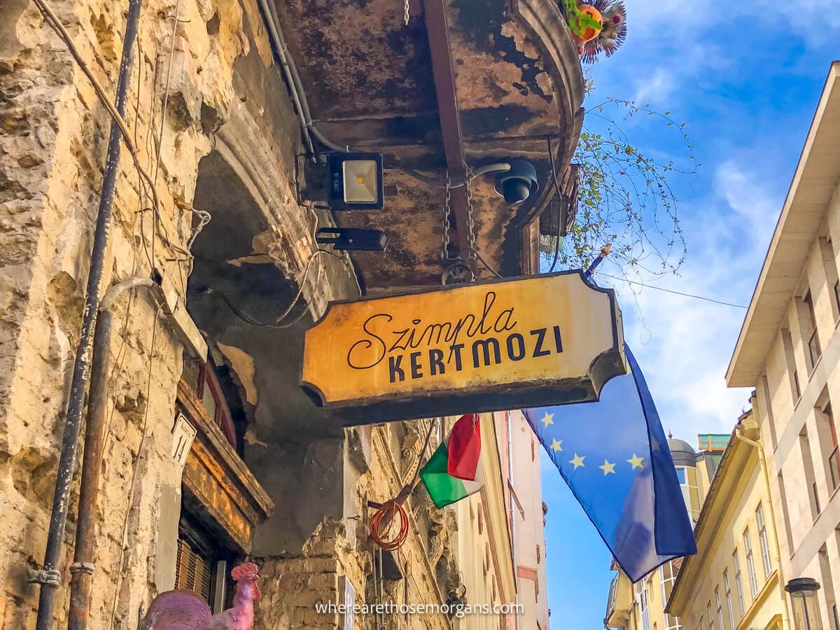 The outside of a tall building in Budapest with a flag flying and a sign for Szimpla Kert ruin bar on a clear sunny day