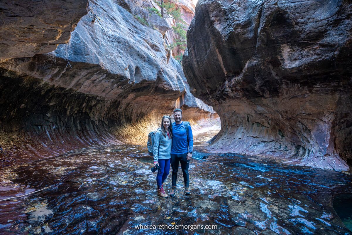 Photo of two hikers stood together inside a tunnel like rock formation called The Subway with orange light bursting in from behind the tunnel