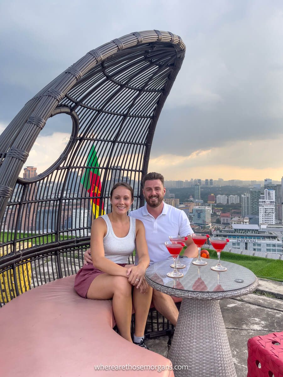 Photo of a couple sat together on a curving bench with a table full of drinks and a rooftop view over a city