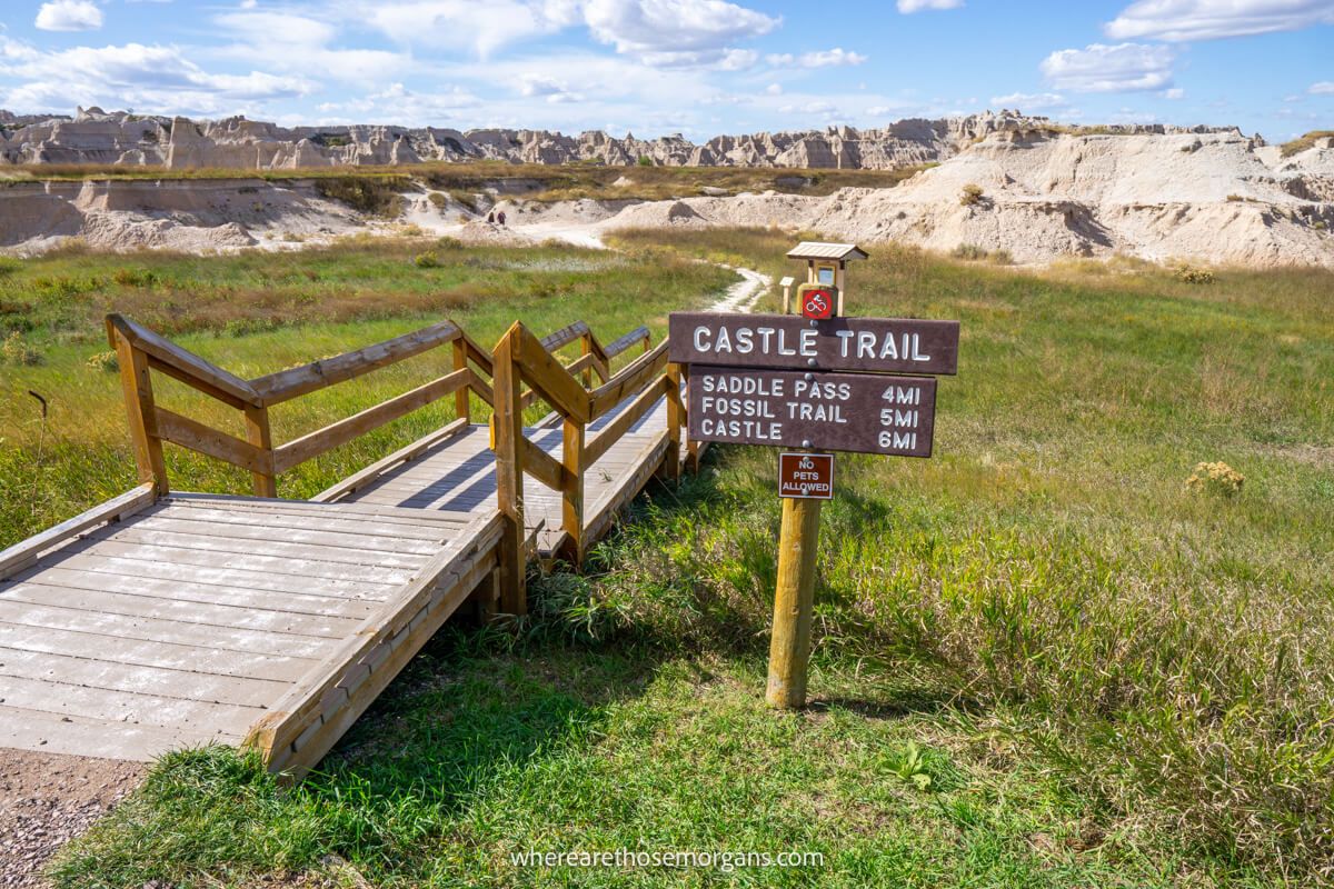 Wooden boardwalk and trail sign leading through grass to rocks and a blue sky