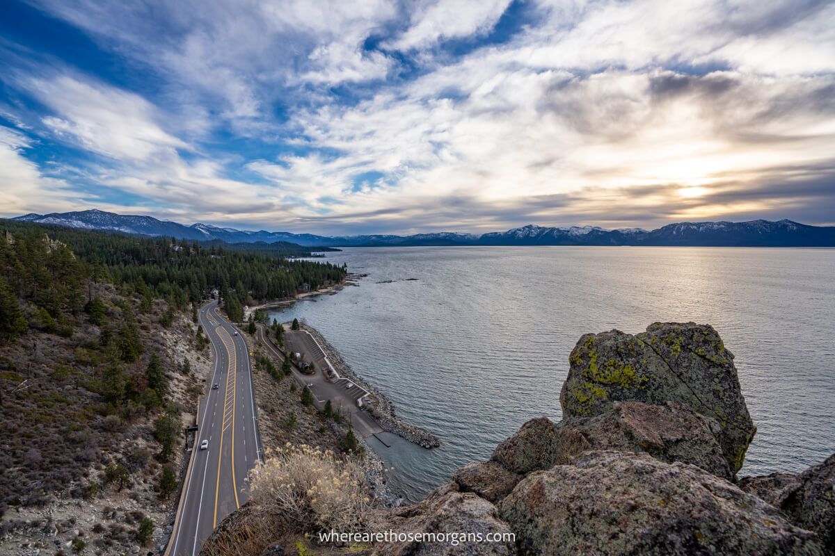 Wide angle photo taken from a rocky summit with boulders overlooking a lakeside road with trees and lots of wispy clouds in the sky at sunset