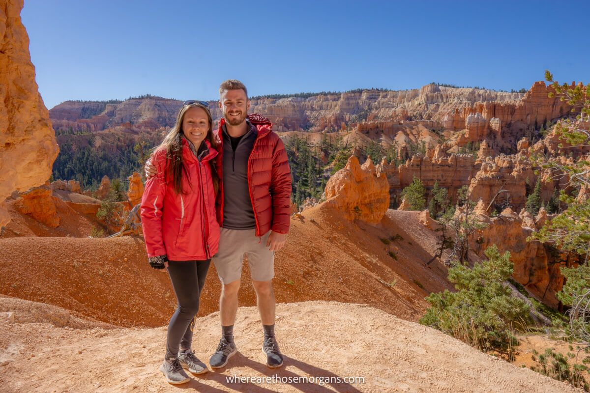 Couple standing together in winter coats on a red rock ledge with hoodoos and cliffs in the background on a sunny day in Bryce Canyon Utah