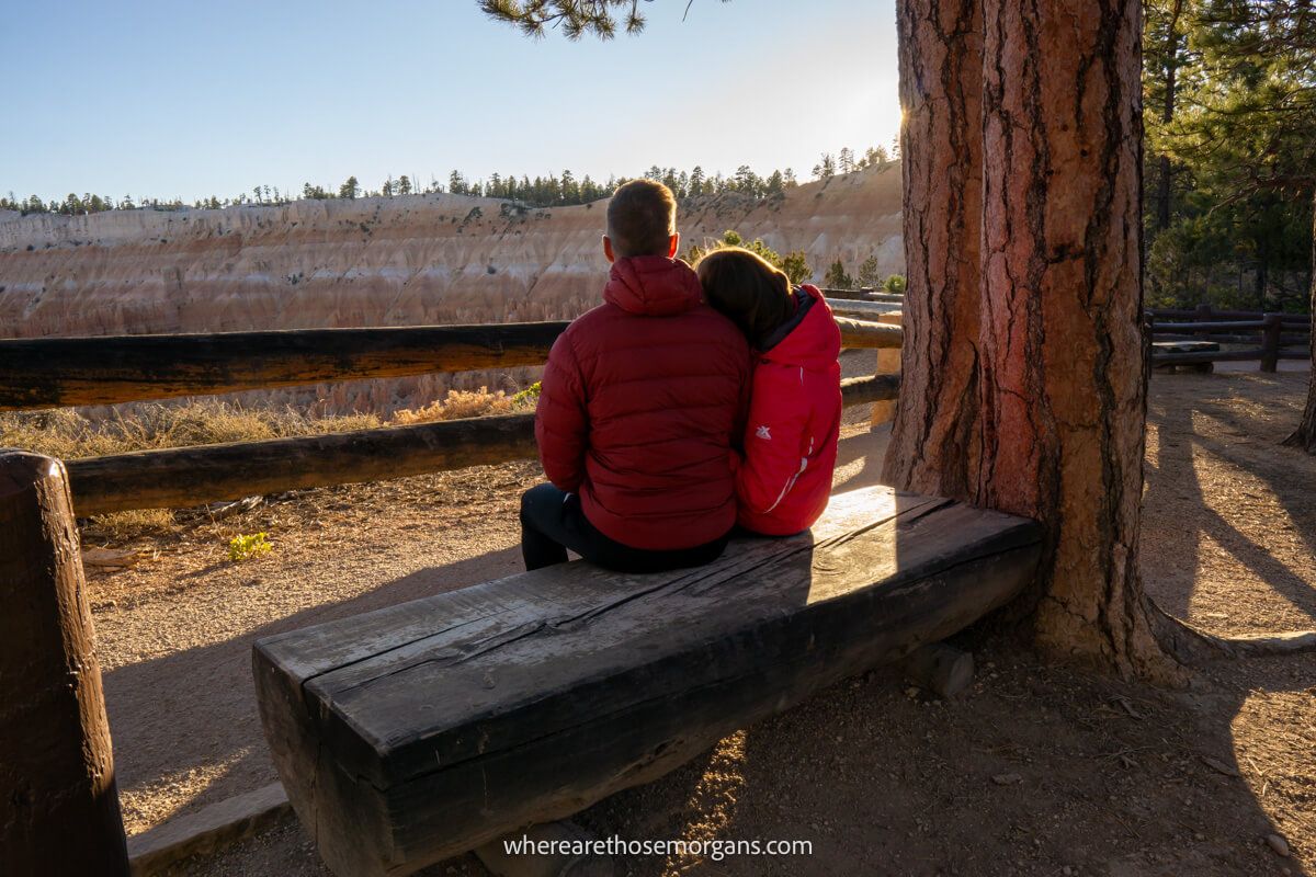 Couple sat on a bench next to a tree at sunset overlooking a sandstone rim in Utah