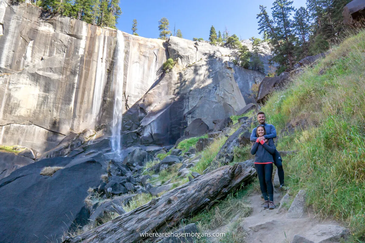 Photo of a couple standing together on a hiking trail near a log on the ground and a thin wispy waterfall cascading over a shelf-like rock formation behind