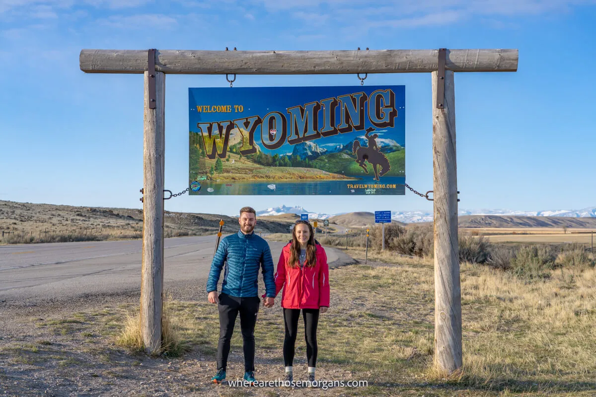 Couple standing together in light winter coats underneath a welcome to Wyoming sign held up by wooden beams