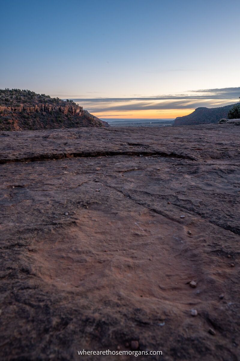 Photo of a dinosaur footprint carved into a flat rock with distant views over red rocks and a soft blue sky at sunset