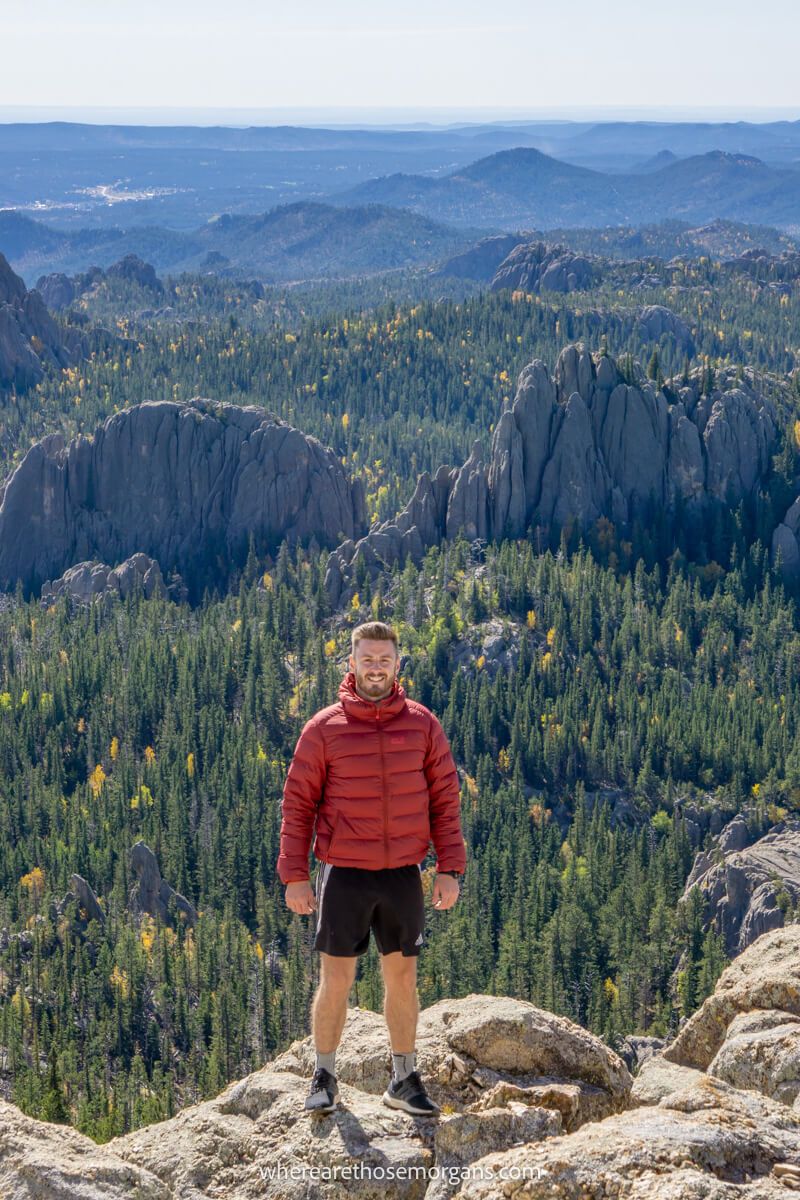 Hiker in a red coat and shorts standing on boulders at the summit of a granite mountain with distant views over forests and granite rocks