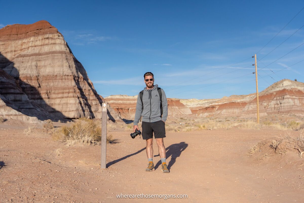 Hiker in grey hoodie and sunglasses holding a camera at the start of Toadstool Hoodoos hiking trail in Utah