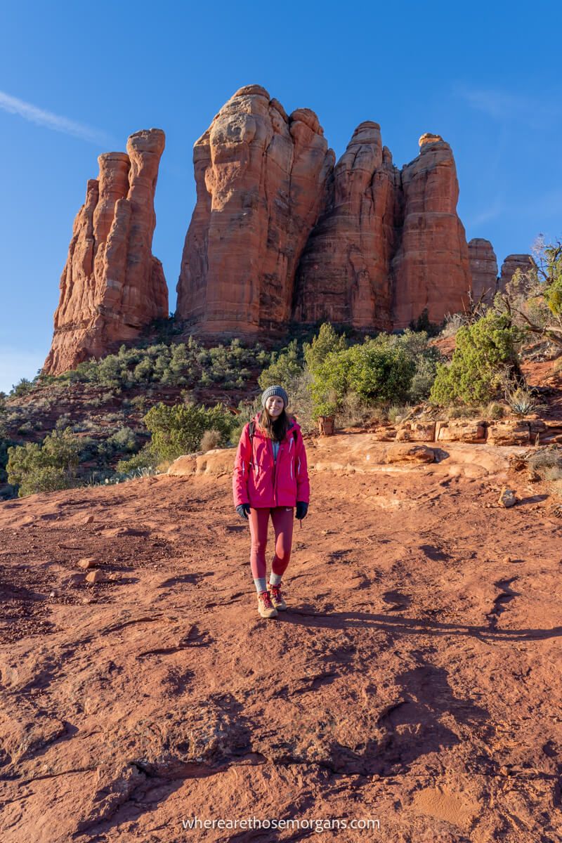 Hiker in winter clothes walking on a sloping red rock landscape with towering sandstone formations behind on a clear day in Sedona AZ
