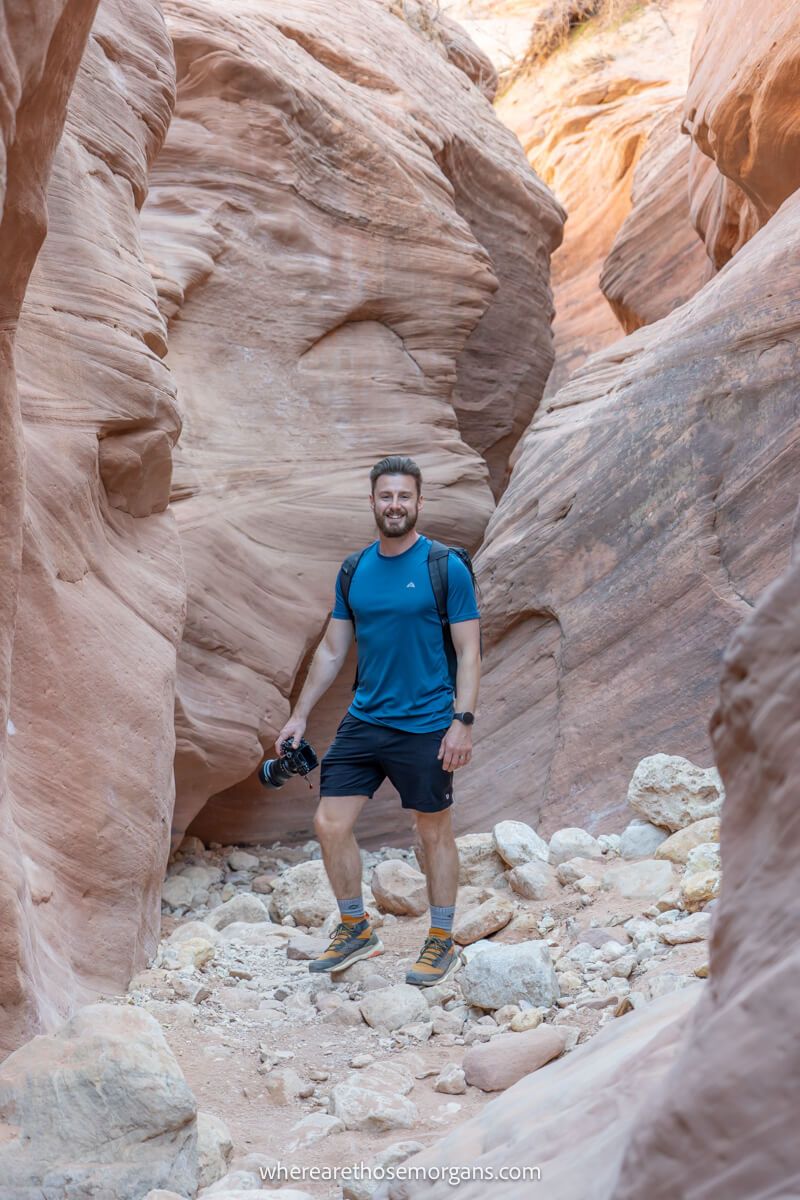 Hiker in blue shirt holding camera and walking through a tall but narrow slot canyon filled with stones