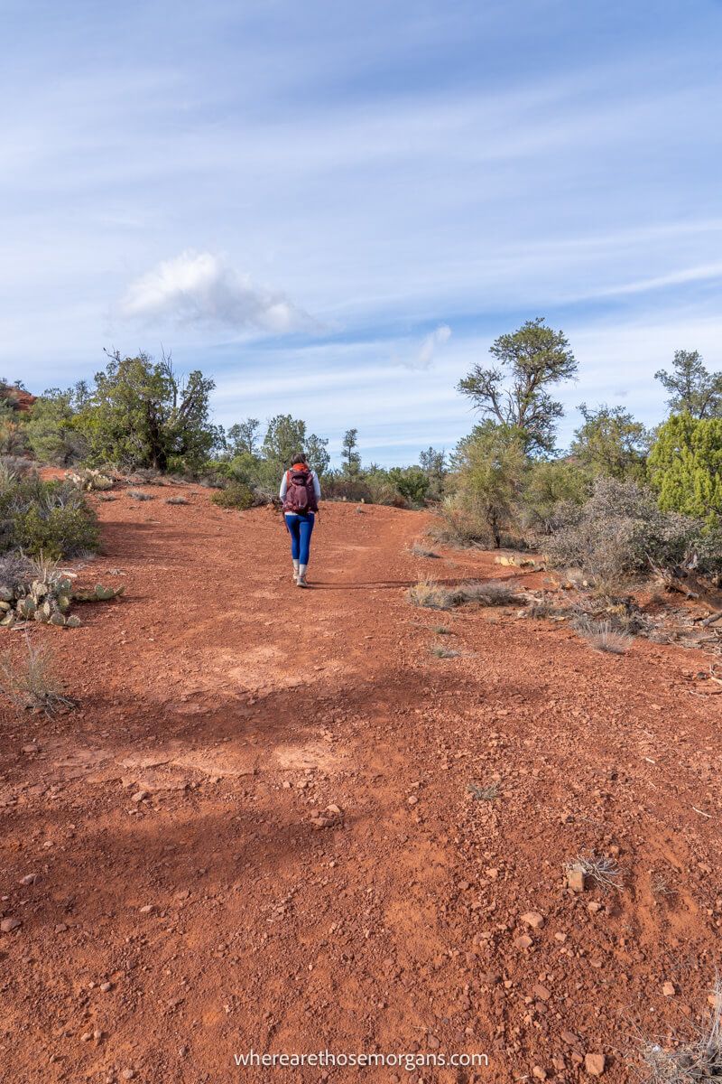 Hiker walking up a gradually inclining red rock trail with trees and a blue sky