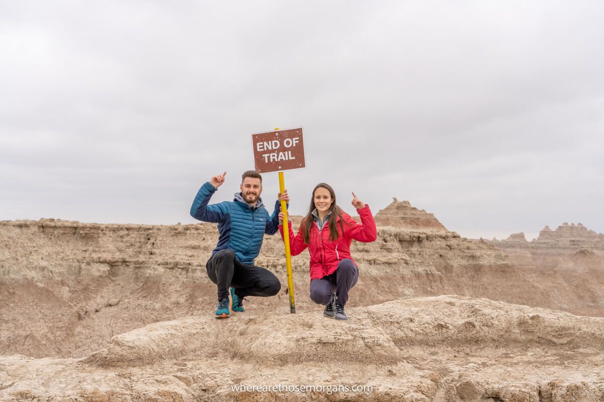 Hikers crouched down for a photo with a sign in Badlands National Park in South Dakota's Black Hills on a cloudy day