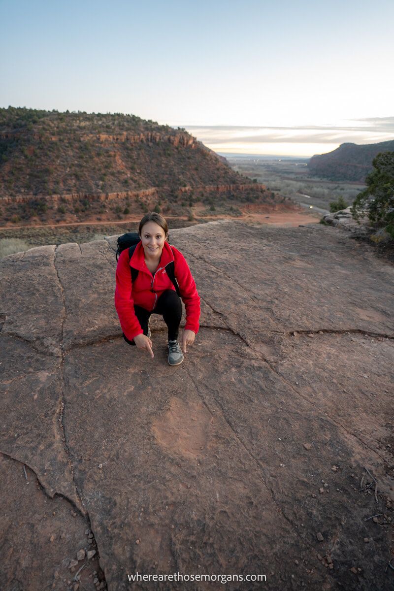 Hiker crouched down on a flat rocky summit pointing at a dinosaur footprint in Utah