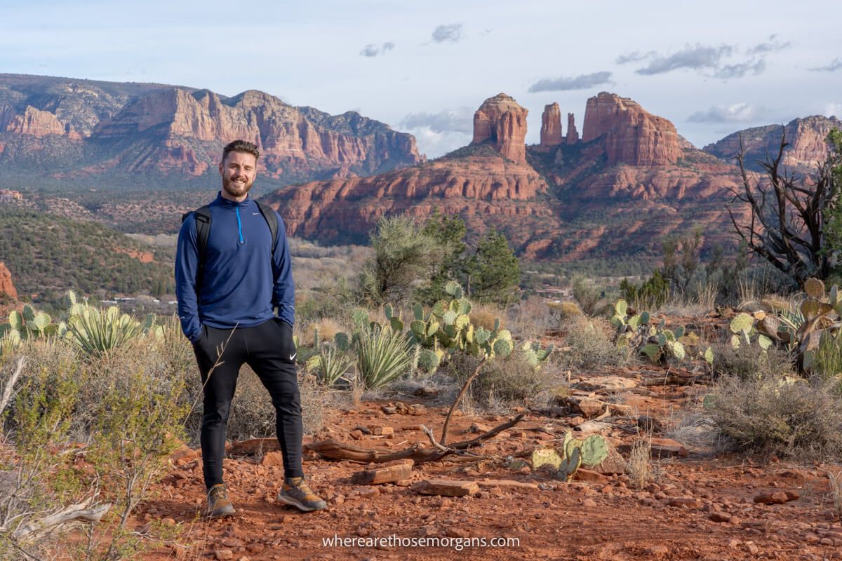 Hiker in black pants and blue top standing for a photo in a desert landscape with red rock formations in the distance