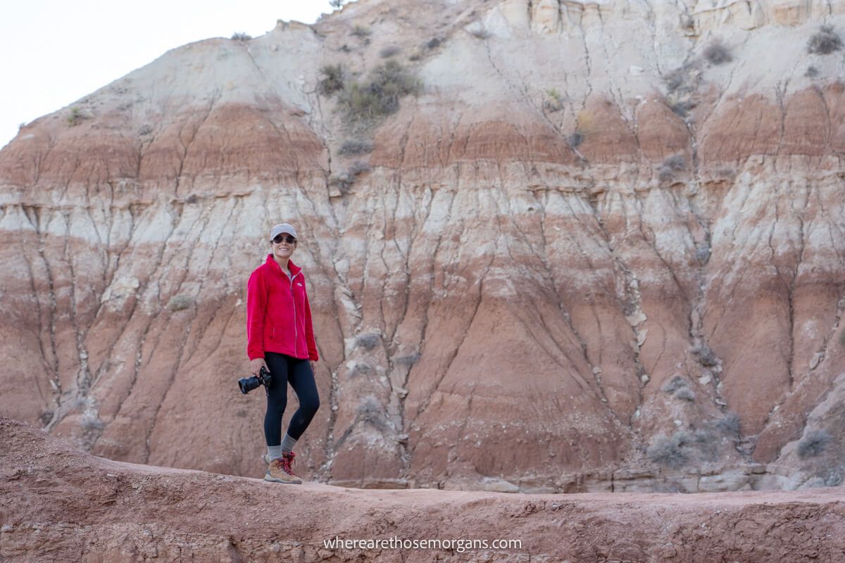 Kristen Morgan wearing black pants and pink coat holding a camera and walking along a rocky ridge with red rock cliffs behind on the Toadstool Hoodoos Trail in Utah