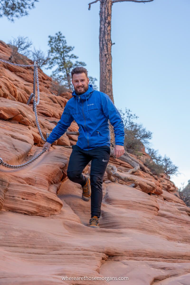 Photo of a hiker in blue coat climbing down a steep rock face holding a metal chain on a cold morning