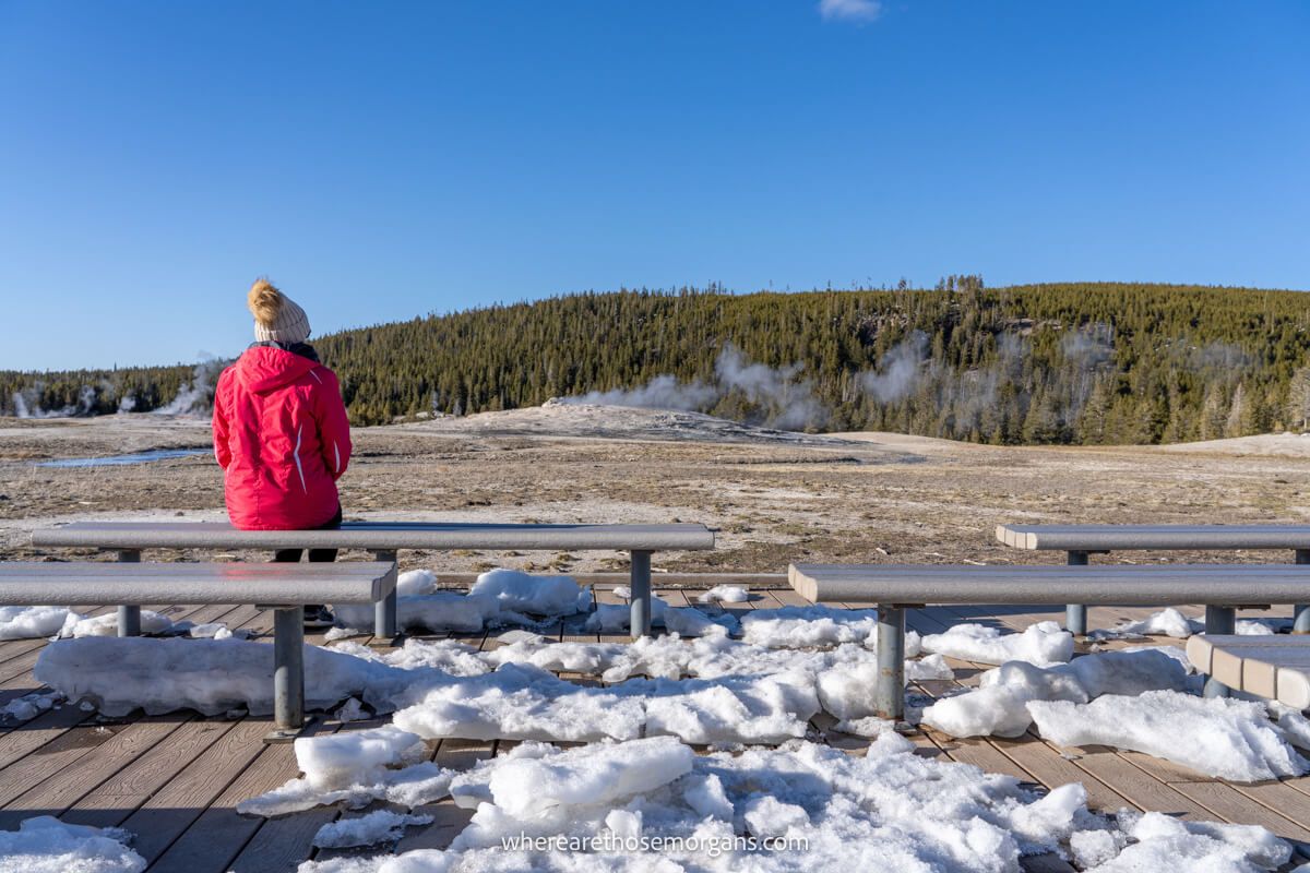 Photo of a person sat alone on curving benches with snow on the ground watching a geyser and waiting for an eruption
