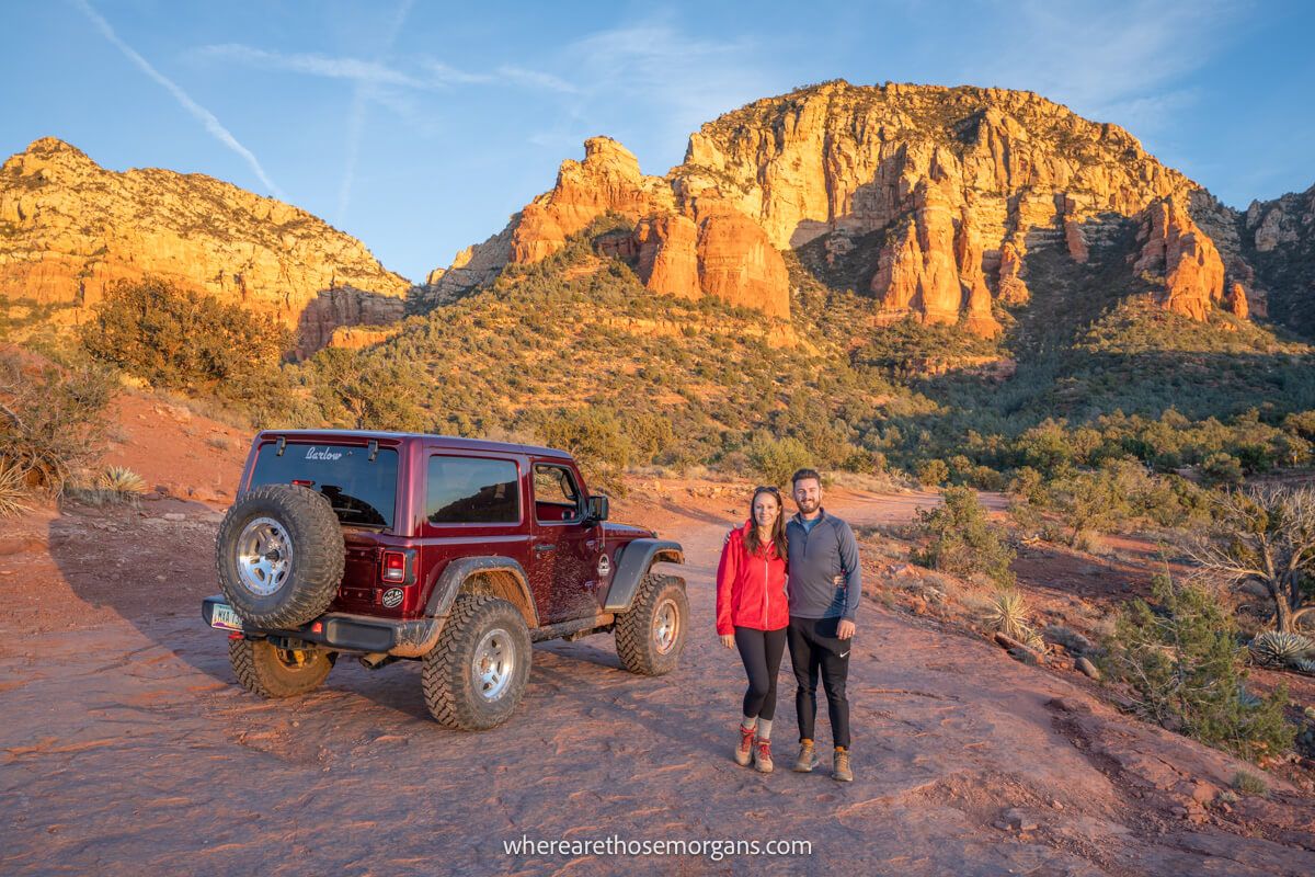 Couple standing together next to a maroon Jeep on a red rock flat table like expanse and towering red cliffs bursting up behind