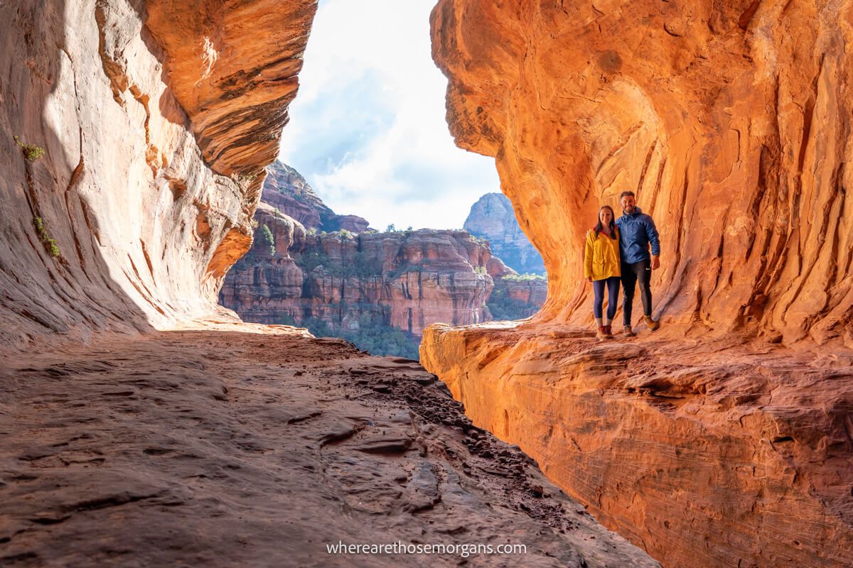 Photo of a couple standing together on one side of a tunnel shaped cave with a hole in the ground and the sky, and views over red rocks through the end of the tunnel formation
