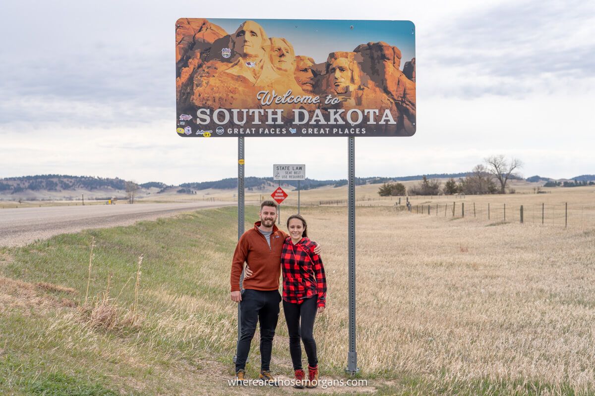 Couple standing together underneath a welcome to South Dakota sign on the side of a road in a wide open landscape