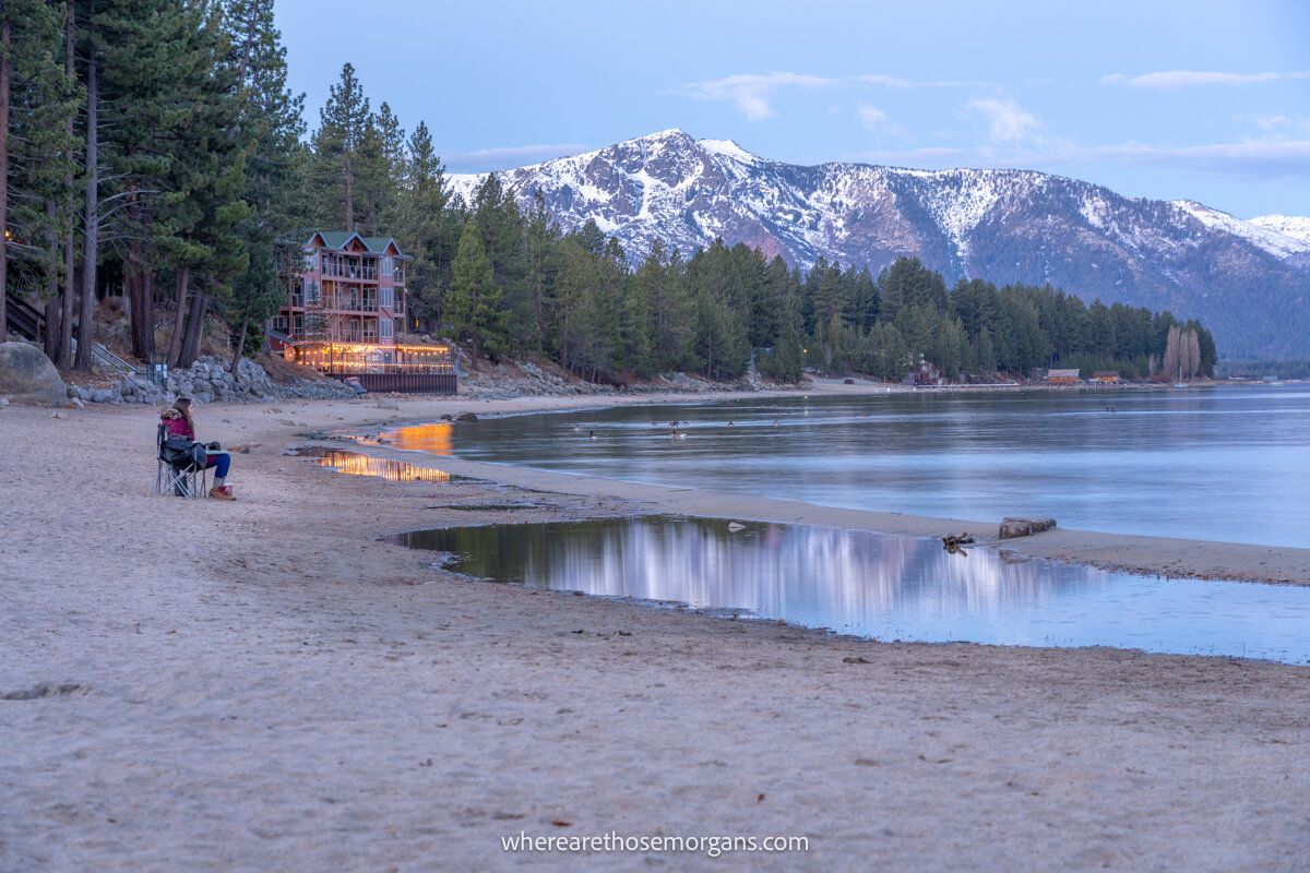 Photographer sat in deck chair on a beach next to a lake waiting for sunrise with mountains and a wooden building in the background
