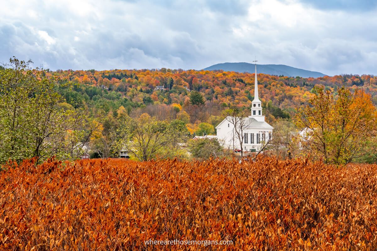 Colorful bushes leading to a white church and hills covered in colorful leaves with clouds in the sky