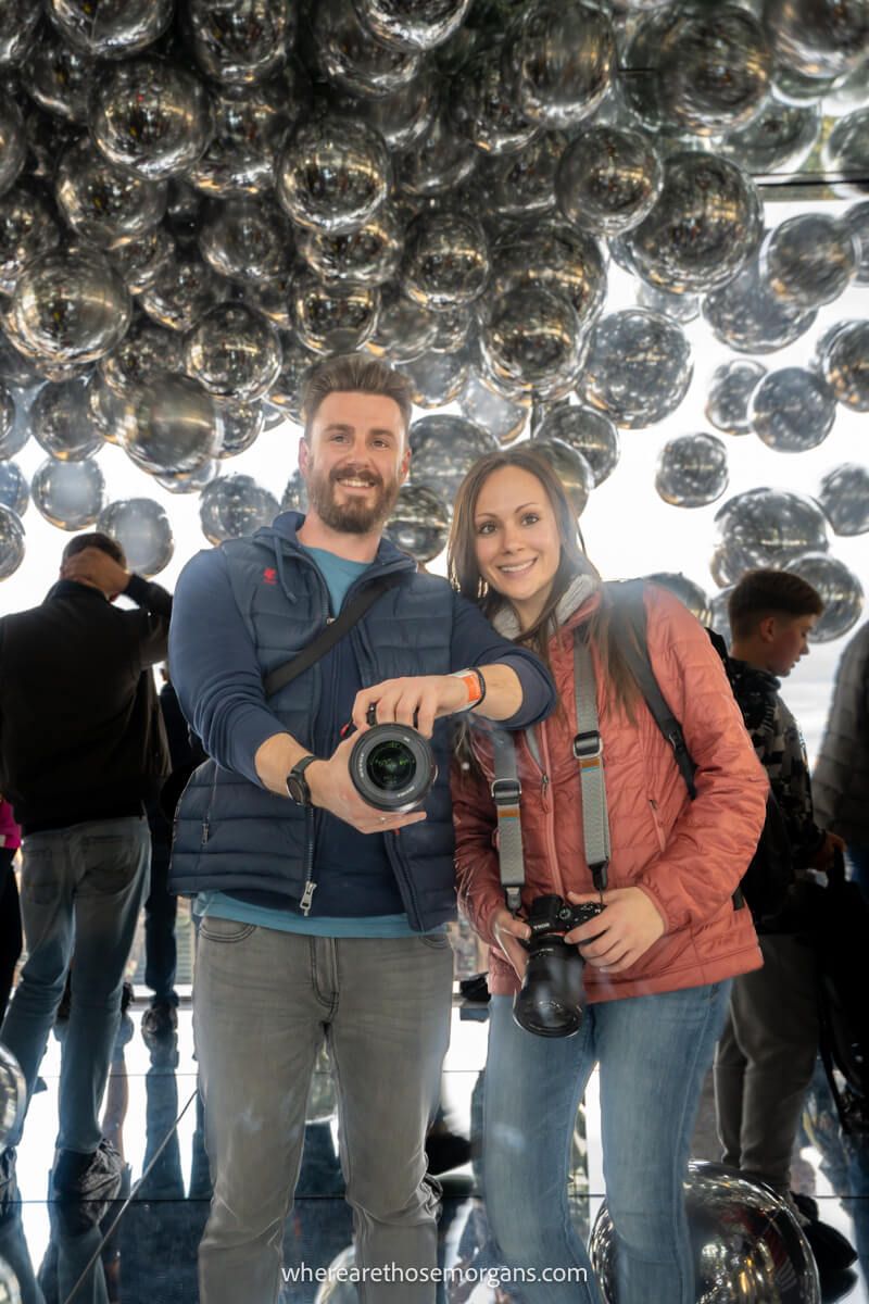 Tourists with cameras standing next to each other taking a photo into a mirror with lots of silver balloons on the ceiling behind