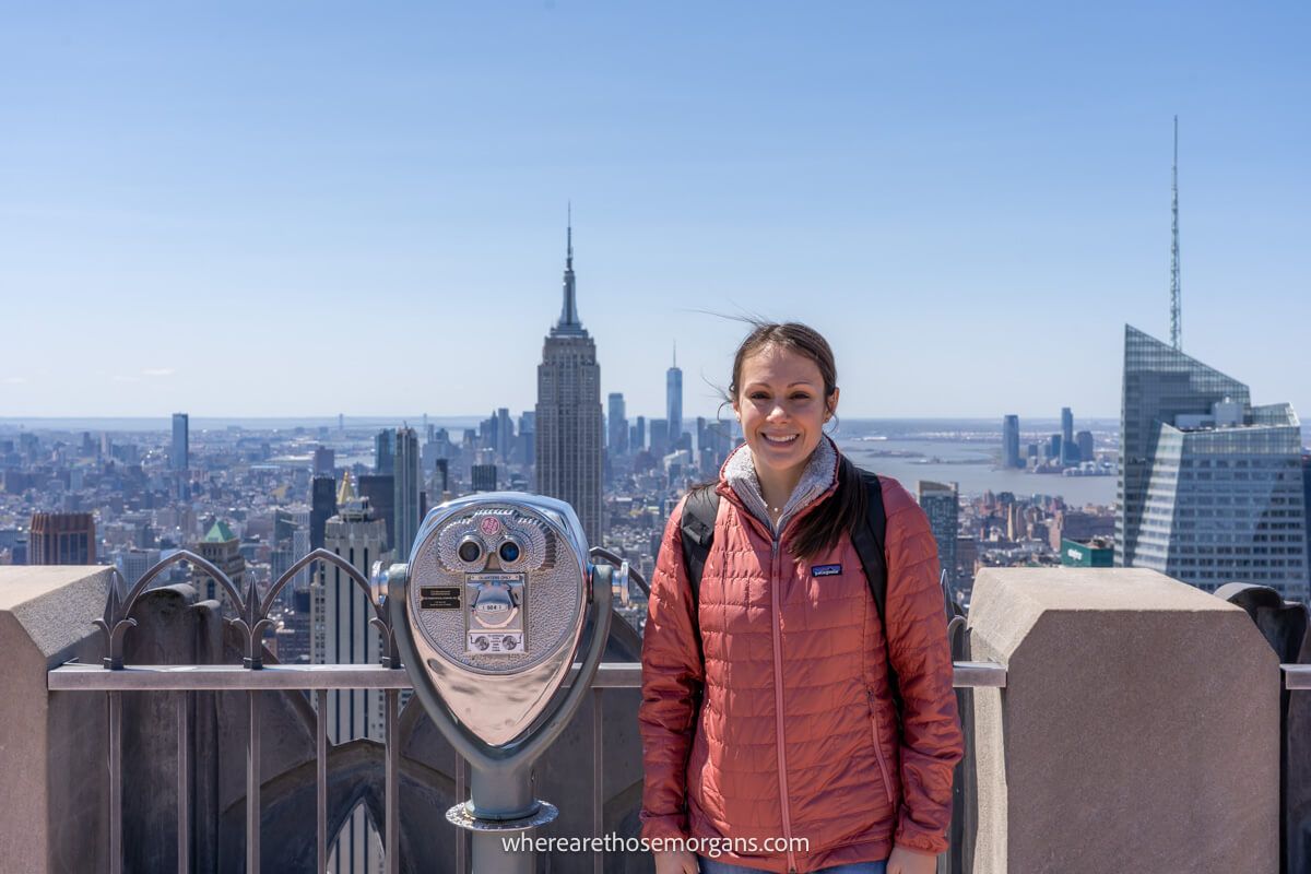 Tourist at Top of the Rock in a red coat next to binoculars overlooking New York City on a clear and sunny day