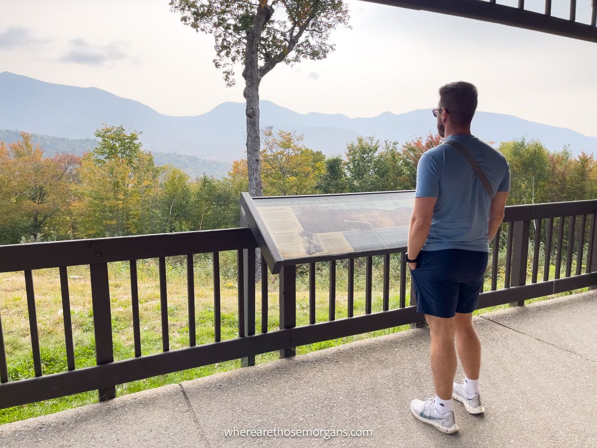 Tourist in shorts and t-shirt with camera at an information board inside a wooden pavilion overlooking a view of trees and hills