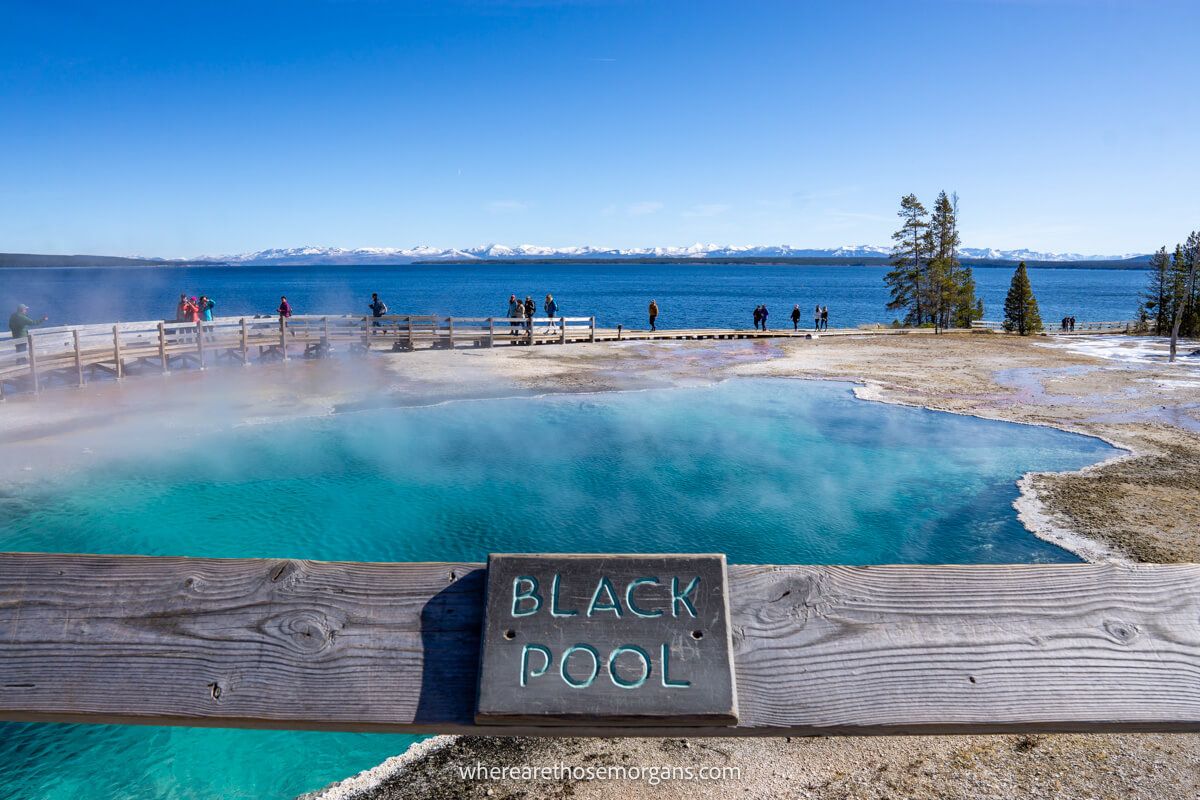 Photo of Black Pool in West Thumb Geyser Basin sign attached to a wooden fence with deep blue got spring behind leading to a large lake and distant mountains