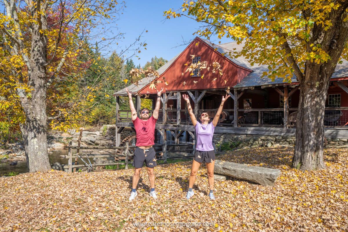 Couple standing next to each other throwing leaves up into the air at the same time with a wooden building behind and trees to the side