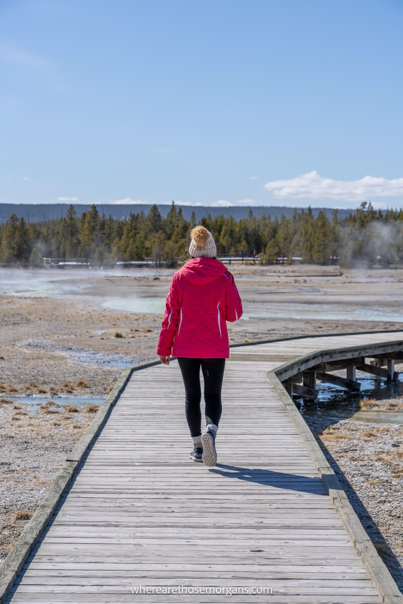Tourist in pink fleece and hat walking alone on a narrow wooden boardwalk through a geyser field on a sunny day