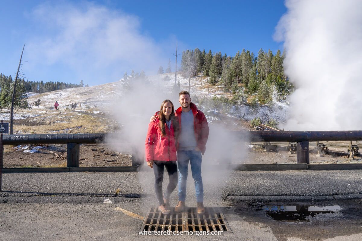 Couple standing together  on top of a vent with steam billowing out and more steam coming out of a hole in the ground behind on a hillside