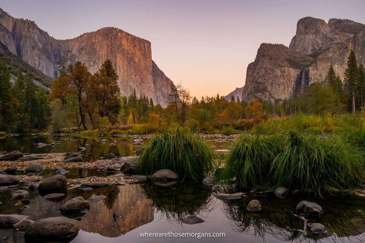 Photo of granite rocks glowing pink at sunrise and green vegetation reflecting in a still river at Valley View in Yosemite